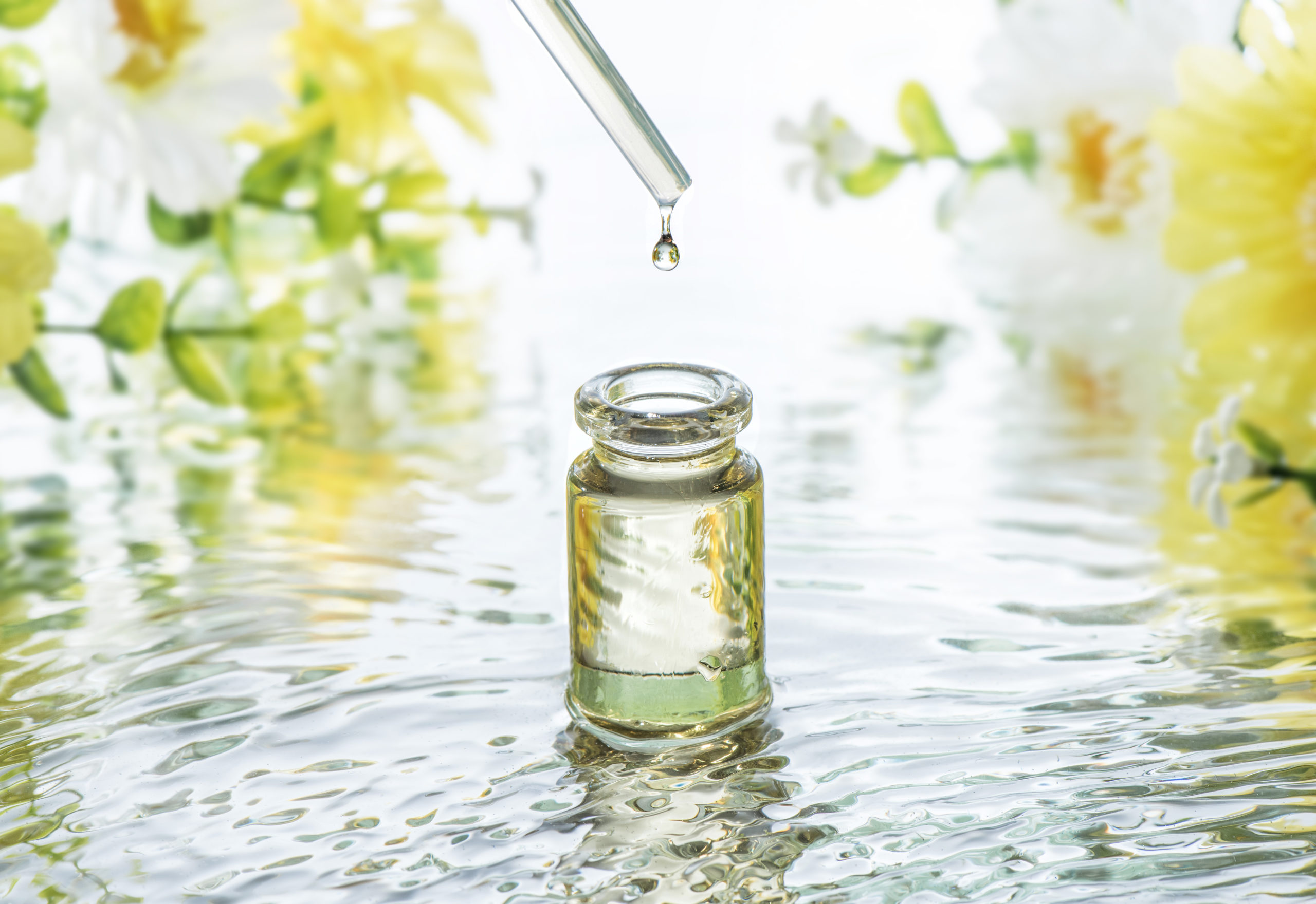 The bottle of Moisturizing cosmetic oil in the water waves on the summer flowers blur background and pipette with oil drop above the bottle